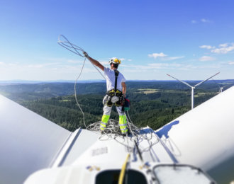 Rear view on Professional rope access technician standing on roof (hub) of wind turbine and pulling rope up. Sun is behind wind turbine.